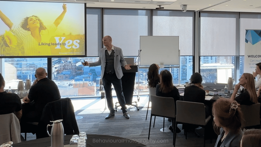 Patrick van der Burght presenting persuasion science to a Melbourne audience, with an image visible on the power point projection of a woman raising her arms in an enthusiastic response.