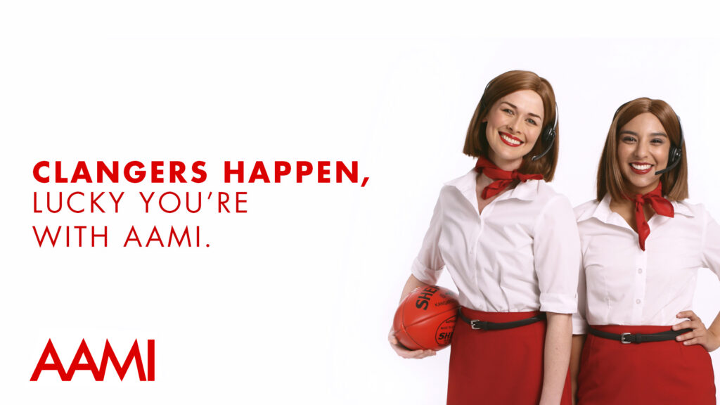 AAMI insurance advertisement featuring two smiling women from different cultural backgrounds in white shirts and red skirts, with one holding an Australian rules football beside the slogan “Clangers happen, lucky you're with AAMI.”